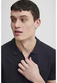 Navy polo shirt featuring a classic collar and a textured fabric. The model's hand is positioned at the neckline, revealing the shirt's fit.