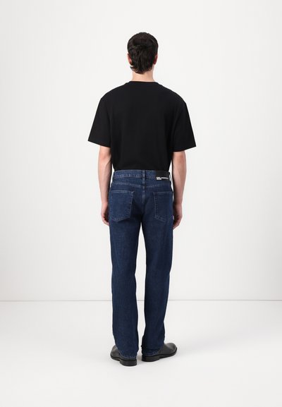 T-shirt noir à manches courtes au style décontracté, associé à un jean en denim bleu foncé avec poches arrière et une étiquette de marque discrète.