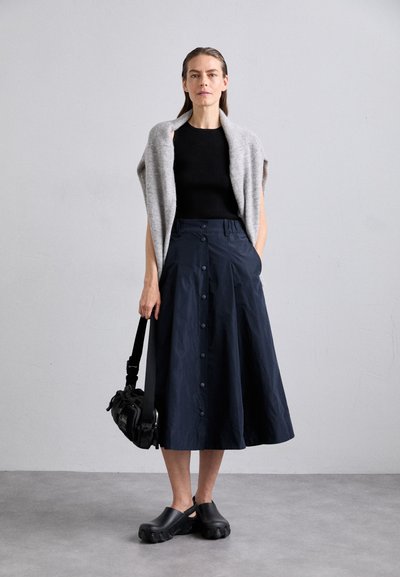 Jupe A-ligne bleu marine avec des détails de boutons, associée à un haut noir ajusté et un châle gris. Des sabots noirs et un sac noir complètent la tenue.