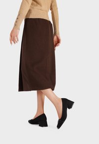 Scarpe nere in suede con tacco quadrato e punta affusolata, adornate con dettagli hardware, abbinate a una gonna midi marrone e un maglione beige.