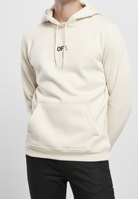 Sweat à capuche beige avec une poche kangourou à l'avant et une capuche à cordon. Présente le texte "OFF." en noir, fabriqué en tissu doux.