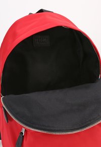 Röd ryggsäck med ett dragkedjefack, svart innertyg och rektangulär form. Har en logotypetikett på insidan. Slät yta.