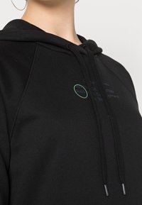 Černá mikina s kapucí, obsahující klokaní kapsu, stahovací šňůrku a tištěné detaily loga v zelené a modré barvě. Vyrobena z hladké, texturované tkaniny.