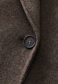Bruine wollen stof met een zachte textuur, voorzien van een donkerbruine knoop met vier gaten, bevestigd met een bruine draad.