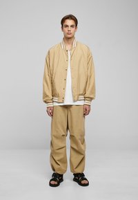 Giubbotto bomber beige con rifiniture a strisce bianche, indossato sopra una camicia bianca. Abbinato a pantaloni beige larghi e sandali neri, in piedi su uno sfondo semplice.