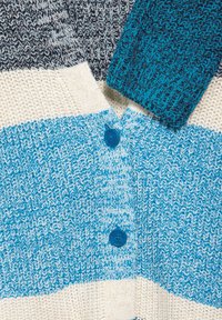 Stickad kofta med horisontella ränder i blått, vitt och marinblått. Två blå knappar och ett texturerat tyg med en mjuk, vävd yta.
