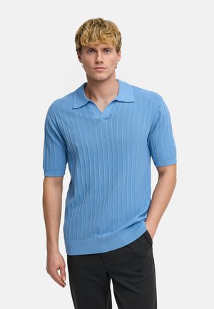 Giovane uomo con capelli biondi che indossa una maglietta polo a maniche corte a coste azzurro chiaro e pantaloni neri, in piedi con una mano in tasca.