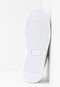 Semelle blanche de chaussure Puma avec un motif clouté carré et des lignes ondulées à l’avant et au talon, appuyée contre un mur d’angle blanc.