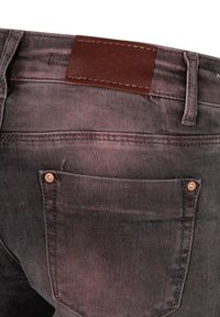 Jeans de mezclilla negra con un parche de cinturón de cuero marrón, presentan remaches de cobre y un bolsillo trasero, mostrando una textura desgastada.
