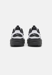 Svartvita träningsskor med en texturerad gummisula, vadderad krage och framträdande varumärkeslogotyp på baksidan. Har en slank, modern design.