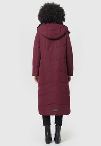 Bordeaux pufferjas met een capuchon en lange lengte, voorzien van gestikte panelen, zijzakken en een logo-detail bij de zoom. Draagt met zwarte broek en laarzen.