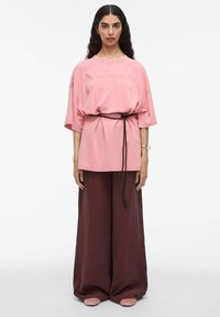 Roza oversize majica s natpisom "ROUGE EDIT" na prednji strani, zategnjena v pasu z črnim trakom, v kombinaciji s širokimi temno burgundskimi hlačami.