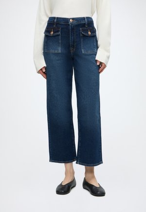 Kvinde iført mørkeblå vidde jeans med frontlommer med klap og sorte flade sko, kombineret med en hvid langærmet top.