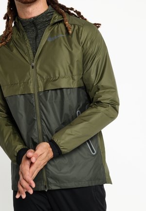 Mężczyzna w oliwkowo-zielonej i ciemnoszarej lekkiej kurtce zapinanej na zamek z bocznymi kieszeniami na zamek, stojący z rękami złożonymi przed sobą.