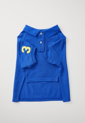 Mėlynas polo marškinėlis, pagamintas iš tekstūruoto medvilnės. Yra apykaklė, trys mygtukai, trumpomis rankovėmis su geltonu skaičiumi 3 kairėje rankovėje.