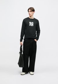 Μαύρο μπλουζάκι με μακριά μανίκια και λογότυπο, φαρδιά μαύρα παντελόνια και εκρού αθλητικά παπούτσια. Το μοντέλο κρατάει μία μαύρη τσάντα με δύο λουριά.