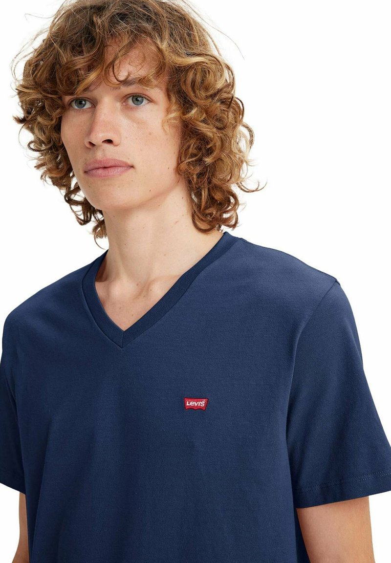 Junge Person mit lockigem Haar, die ein dunkelblaues T-Shirt mit V-Ausschnitt und einem kleinen roten Levi's-Logo auf der Brust trägt.