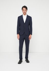 Ναυτικό κοστούμι με slim fit, σε μονόχαρη σχεδίαση, με τρύπες και δύο κουμπιά. Συνοδεύεται από λευκό πουκάμισο και μαύρα παπούτσια.