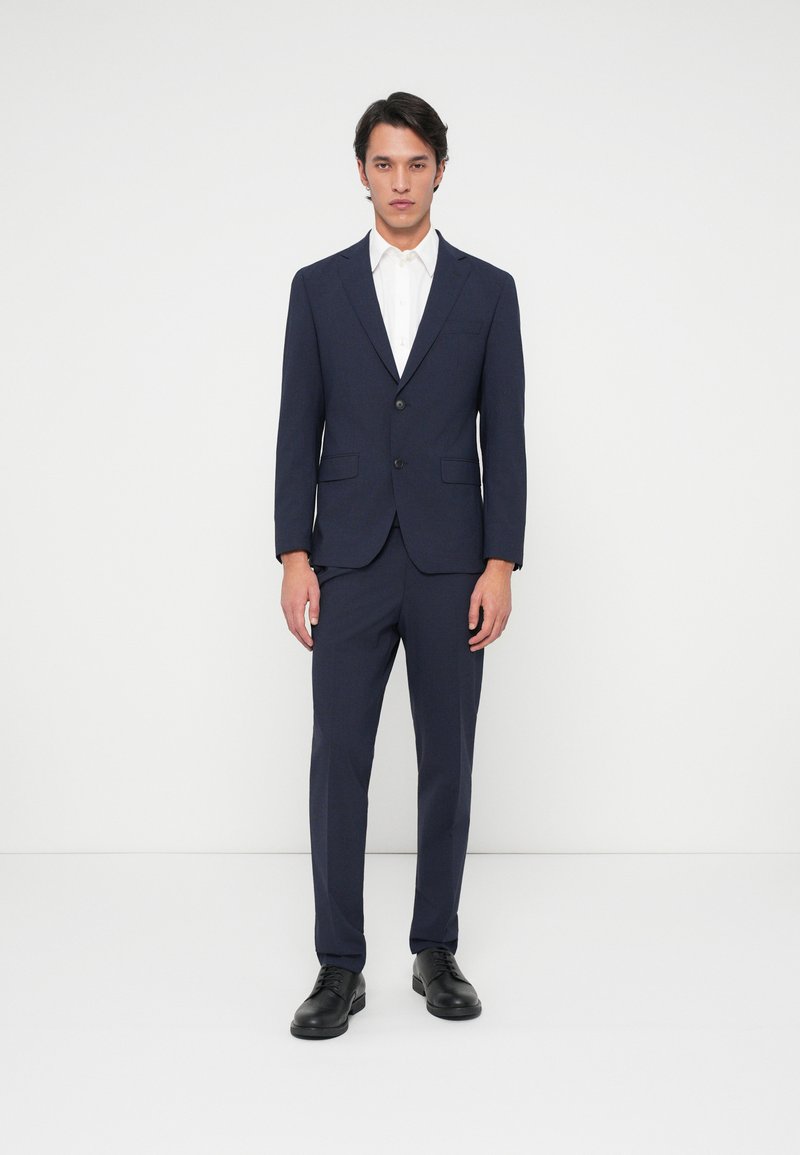 Ναυτικό κοστούμι με slim fit, σε μονόχαρη σχεδίαση, με τρύπες και δύο κουμπιά. Συνοδεύεται από λευκό πουκάμισο και μαύρα παπούτσια.