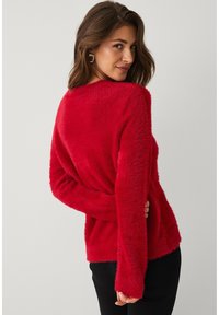 Rode, pluizige trui met een zachte textuur, losse pasvorm en lange mouwen. Het heeft een ronde halslijn en wordt gecombineerd met een zwarte broek.