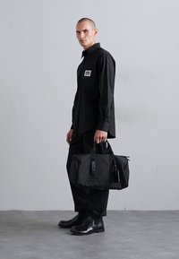 Sac de sport noir au fini texturé, doté de poignées doubles, d'une fermeture zippée et d'une étiquette logo. Porté par un mannequin en tenue noire.