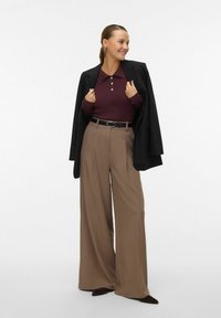 Pantaloni larghi di un marrone scuro, polo a coste color bordeaux con pattina di bottoni, blazer nero adagiato sulle spalle e cintura nera con fibbia rotonda.
