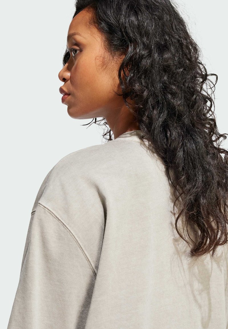 Sudadera beige de corte holgado, escote redondo y costuras caídas en los hombros. El cabello es rizado y cae sobre un hombro.