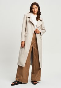 Cappotto in misto lana grigio chiaro con rever a punte, chiusura a un bottone e tasche laterali, indossato sopra un top bianco e pantaloni a gamba larga marroni.