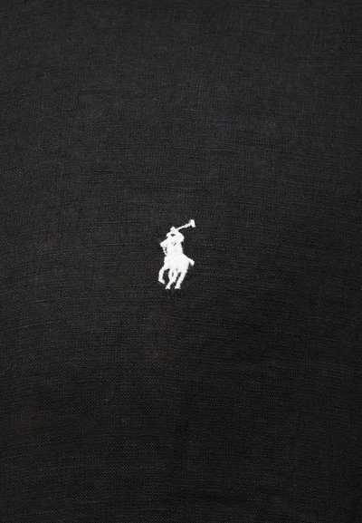 Logotipo blanco bordado de un jugador de polo a caballo que balancea un mazo sobre una tela negra texturizada.