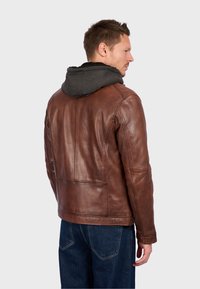 Man in een bruine leren jas met een grijze hoodie eronder en blauwe spijkerbroek, staand met zijn gezicht naar rechts tegen een effen witte achtergrond.