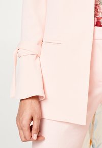 Dłoń kobiety obok jasnoróżowej marynarki z wiązanym detalem przy rękawach oraz dopasowanych spodni, częściowo widoczna kwiatowa koszula.