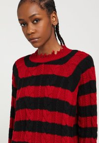 Červený a čierny pruhovaný pletený sveter s kabelovým vzorom, so zničeným okrúhlym výstrihom a dlhými rukávmi. Mäkka textúra a voľný strih.