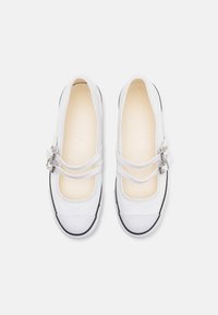 Zapatillas Mary Jane de lona blanca con correas de doble hebilla y puntas de goma, vistas desde arriba sobre un fondo blanco.