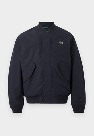 Giubbotto bomber blu navy con collo, polsini e orlo a costine, tasche frontali con patta e piccolo logo coccodrillo verde sul petto a sinistra.