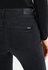 Černé džíny s hladkou texturou, vybavené zadní kapsou a koženým páskem s logem vyraženým "A|X."