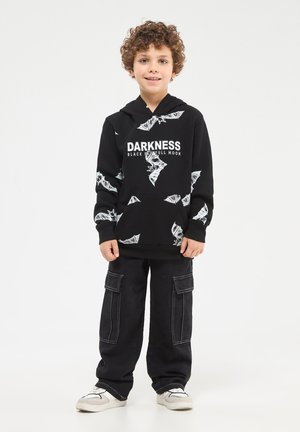 Felpa nera con grafiche di pipistrelli bianchi e testo "DARKNESS BLACK SKULL FULL MOON." Abbinata a pantaloni cargo neri e scarpe da ginnastica bianche.