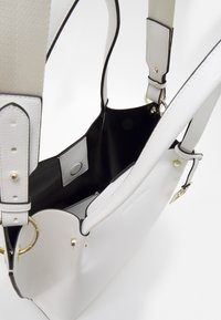 Bílá kůže kabelka s hladkým povrchem, černým interiérem, nastavovacími popruhy, zlatými akcenty a detailním zapínáním na cvok.