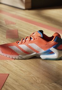 Scarpa sportiva arancione con strisce bianche e accenti blu su pavimento in legno con linee del campo arancioni.