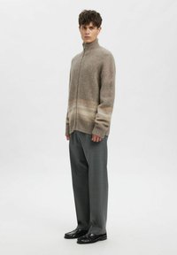 Beige genser med glidelås og høy krage, med et gradientmønster i lysere toner. Brukt over grå bukser og svarte sko.