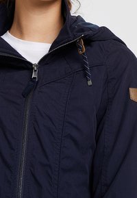 Granatowa kurtka z zamkiem błyskawicznym z przodu, kapturem i sznurkiem ściągającym. Posiada teksturowany materiał i małą brązową skórzaną naszywkę na ramieniu.