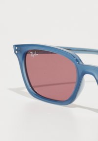 Gafas de sol de plástico azul con grandes monturas angulares y lentes de color rosa oscuro. Presenta el logo de Ray-Ban en la lente superior izquierda.