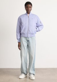 Ljubičasta bomber jakna s prednjim patentnim zatvaračem, rebrastim naramenicama i bočnim džepovima, uparena s svijetloplavim širokim hlačama i bijelim tenisicama.