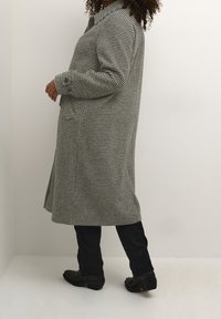 Rutigt kappa i svart och vitt hounds-tooth mönster, med krage, långa ärmar och sidofickor, som bärs tillsammans med mörka jeans och boots.