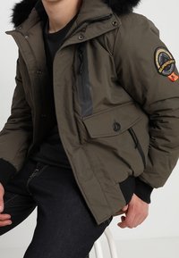 Olivengrøn puffjakke med sort pelsforet hætte, justerbare manchetter, forlommer og et mærke på ærmet.