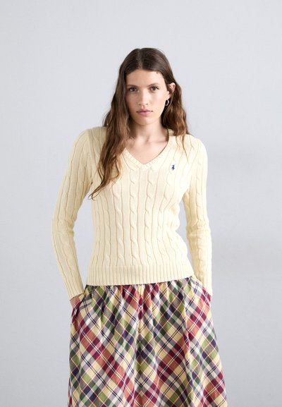 Cable-knit, V-hals sweater i lys gul; tætsiddende silhuet. Matcher en ternet, fuld, farverig nederdel med røde, grønne og beige tern.