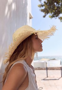 Jeune femme dans un haut sans manches blanc portant un chapeau de paille à larges bords, debout devant un mur blanc avec une vue ensoleillée sur la mer en arrière-plan.
