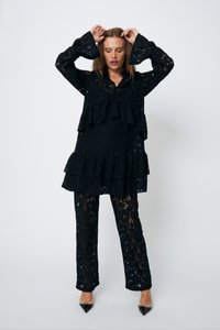 Completo in pizzo nero con un top a peplum a strati e maniche a campana, abbinato a pantaloni attillati. Tessuto trasparente con motivi floreali, indossato con tacchi a punta.