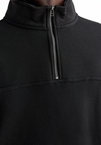 Felpa nera con zip e colletto alto, morbida al tatto, con una zip argentata. Presenta una cucitura orizzontale sul petto per un tocco di dettaglio.