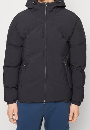Persona che indossa un giubbotto imbottito nero con cappuccio e tasche con zip, abbinato a pantaloni blu, in piedi contro uno sfondo neutro.