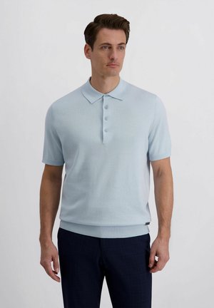 Mann trägt hellblaues kurzärmeliges Polohemd mit Knöpfen und dunkle Hose, steht vor schlichtem weißem Hintergrund.